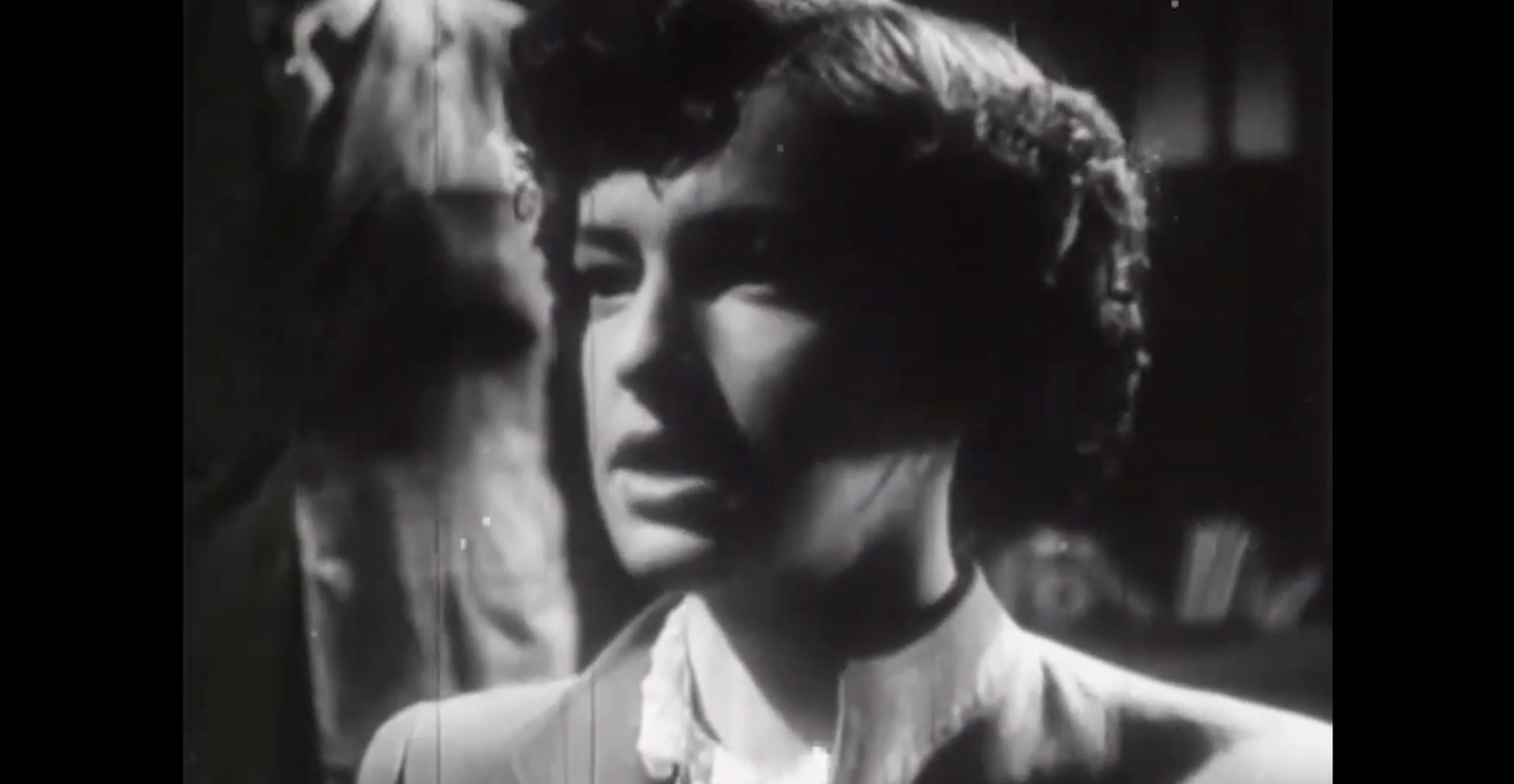 Um close em preto e branco de uma mulher com cabelo curto e ondulado, vestindo uma blusa com gola. A imagem parece ser de um filme antigo, com grãos visíveis e alto contraste, dando um visual cinematográfico clássico. A mulher tem uma expressão séria, ligeiramente angustiada, olhando para a esquerda fora da câmera.